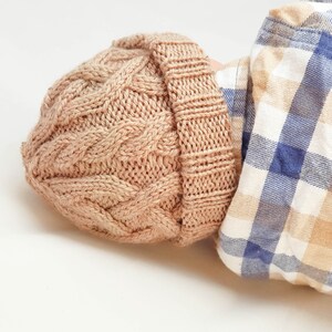 May include: A light brown cable-knit beanie hat. The hat is made of a soft, textured yarn and features a folded brim. The hat is shown with a blue and white plaid shirt.