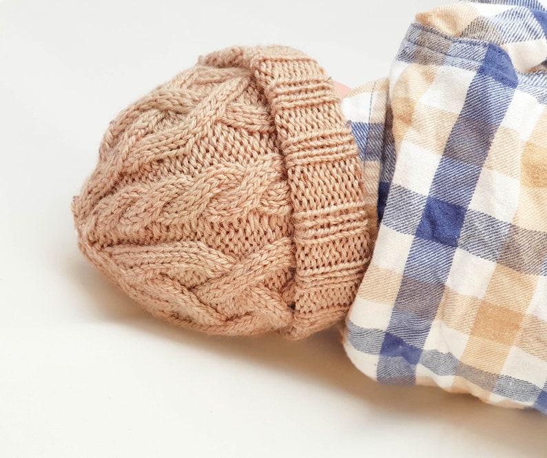 May include: A light brown cable-knit beanie hat. The hat is made of a soft, textured yarn and features a folded brim. The hat is shown with a blue and white plaid shirt.