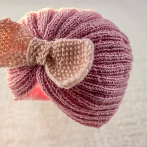 May include: A pink knitted hat with a large pink bow. The hat is made of a soft, textured yarn.