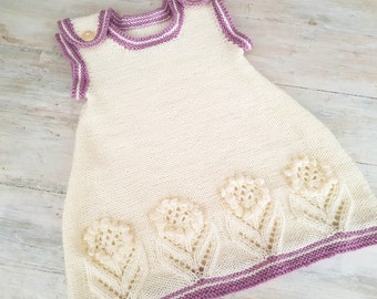 May include: A white knitted baby dress with a purple trim and a delicate floral design around the bottom.