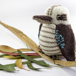 May include: A knitted toy bird with brown and white stripes, blue accents, and a brown wing. The bird is perched on a branch with dried leaves.