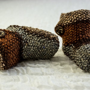 May include: A pair of brown and gray knitted baby booties with a button on each. The booties are made of a soft, textured yarn.