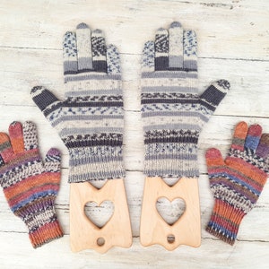 May include: Two pairs of knitted gloves with colorful patterns, one pair is grey and blue, the other is orange and purple. The gloves are displayed on wooden heart-shaped holders.