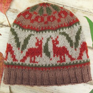 May include: A brown, red, and green knitted hat with a fox pattern. The hat has a brown brim and a white background with red foxes and green leaves.