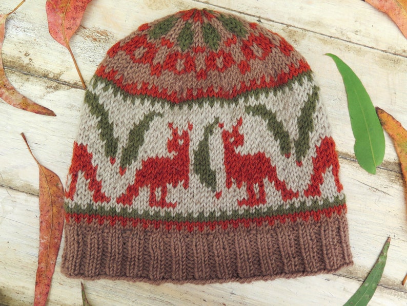 May include: A brown, red, and green knitted hat with a fox pattern. The hat has a brown brim and a white background with red foxes and green leaves.