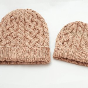 May include: Two knitted cable-knit beanies in a light brown color. The larger beanie has a folded brim and a textured cable pattern across the crown. The smaller beanie has a similar design, suitable for a child or a smaller head.