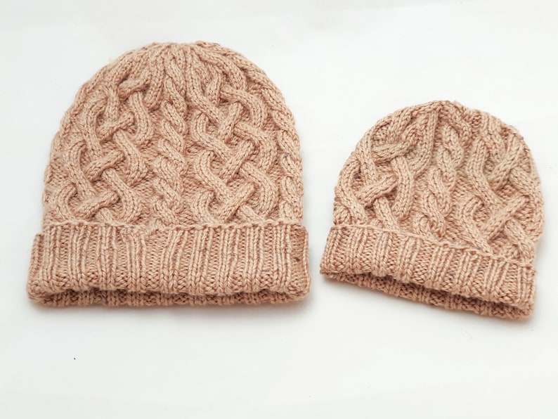 May include: Two knitted cable-knit beanies in a light brown color. The larger beanie has a folded brim and a textured cable pattern across the crown. The smaller beanie has a similar design, suitable for a child or a smaller head.