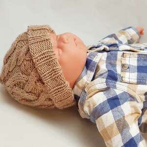 May include: A tan cable-knit baby hat. The hat is made of a soft, textured yarn and features a classic cable-knit design. The hat is shown on a baby doll wearing a blue and tan plaid shirt.
