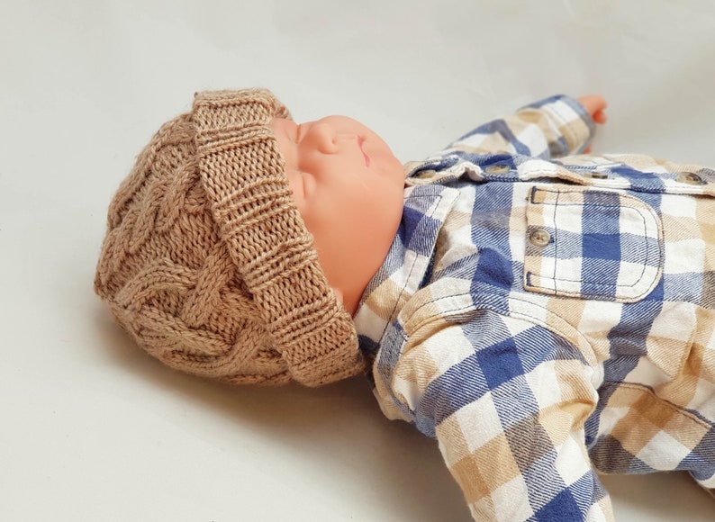 May include: A tan cable-knit baby hat. The hat is made of a soft, textured yarn and features a classic cable-knit design. The hat is shown on a baby doll wearing a blue and tan plaid shirt.