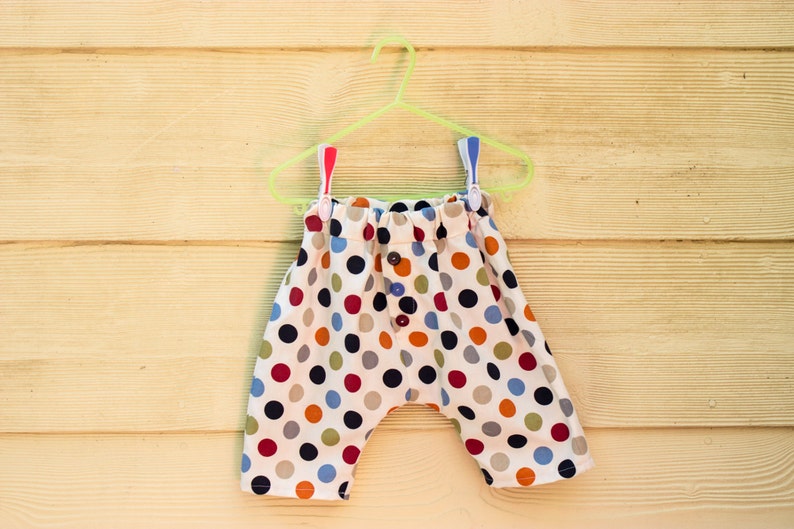 May include: Polka dot patterned pants with a white background and colorful dots. The pants are hanging on a green plastic hanger.