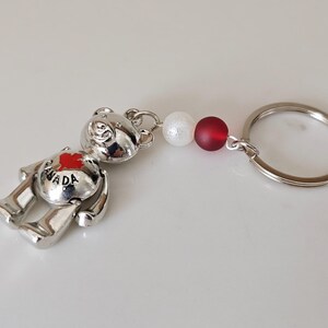 May include: Silver bear keyring with a red maple leaf on its chest that says "CANADA". The keyring has a white and red bead attached to a silver ring.