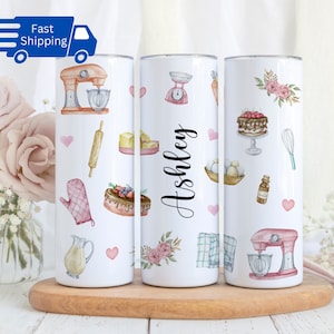 May include: Three white stainless steel tumblers with a baking theme. The tumblers feature a variety of baking-related images, including a mixer, rolling pin, cake, and a whisk. The tumblers are personalized with the name "Ashley".