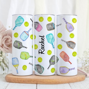 Pu&ograve; includere: Tre bicchieri bianchi con un motivo di racchette da pickleball colorate e palline verdi. Il bicchiere centrale ha il nome "Rachel" scritto su di esso.