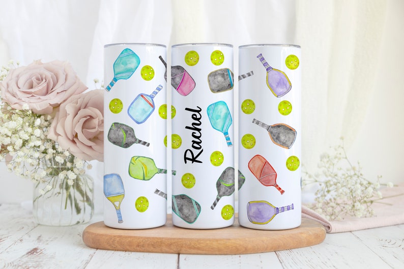 Pu&ograve; includere: Tre bicchieri bianchi con un motivo di racchette da pickleball colorate e palline verdi. Il bicchiere centrale ha il nome "Rachel" scritto su di esso.