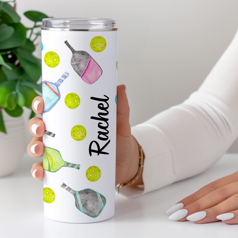 Pu&ograve; includere: Un bicchiere in acciaio inossidabile bianco con un design ad acquerello di racchette e palline da pickleball. Il bicchiere ha il nome "Rachel" stampato su di esso.