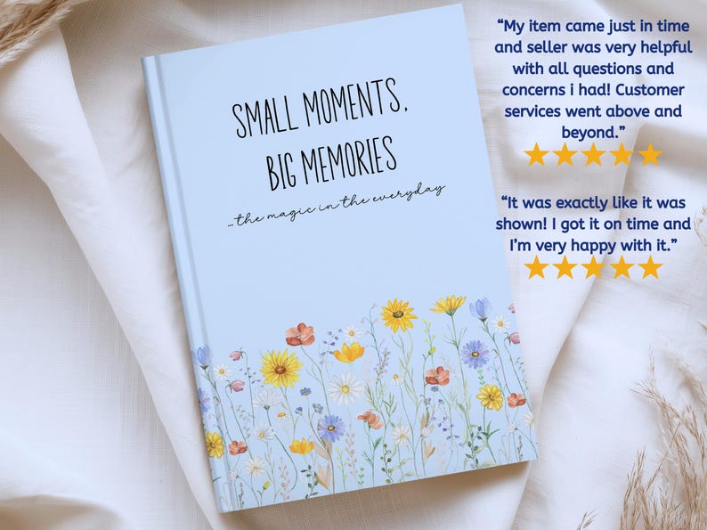May include: A light blue hardcover journal featuring a floral print at the bottom with yellow, orange, and blue wildflowers. The text on the cover reads: SMALL MOMENTS, BIG MEMORIES, the magic in the everyday. The book has a clean, matte finish.