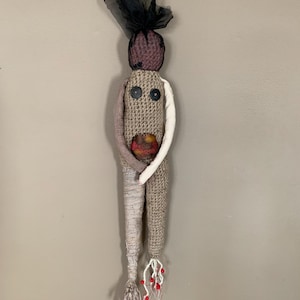 May include: A handmade fabric doll with a textured, beige body and a purple head. The doll has two black button eyes, a white arm, and a colourful centre. The doll's legs are wrapped in fabric, with red bead accents.