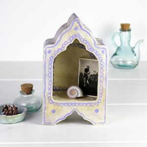 Puede incluir: Una caja de sombras decorativa, hecha a mano, de color blanquecino y lavanda, con una abertura arqueada. En el interior, se ven una fotografía en blanco y negro y una concha. Una pequeña botella de vidrio y un cuenco turquesa con una piña se encuentran en primer plano.