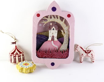 Diorama de una casa en capas hecho a mano sobre una colina y un árbol en tonos rosa, morado y ocre.