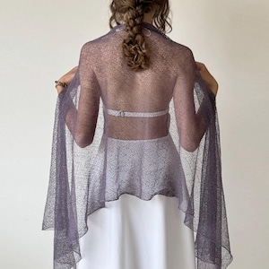 May include: A sheer, lavender-grey shawl with long, flowing sleeves. The shawl is draped over a white dress, creating a layered effect. The fabric has a delicate, dotted texture, adding visual interest to the garment.