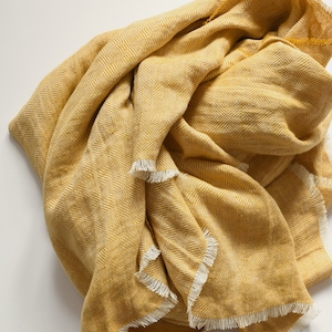 Pode incluir: Um cobertor amarelo dobrado com um padrão de losangos e bordas com franjas. O cobertor é de uma cor mostarda quente e parece macio. A franja é de uma cor branco-sujo contrastante.