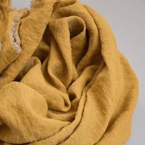 May include: A mustard yellow linen scarf with a fringed edge. The scarf is folded and draped in a way that shows the texture of the fabric.
