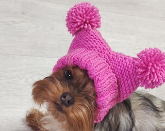 May include: A bright pink knitted hat for a dog, featuring a ribbed brim and two large, round pom-poms attached to the top corners. The hat is crafted from thick yarn with a visible knit texture, designed to fit snugly over the head of a small dog.