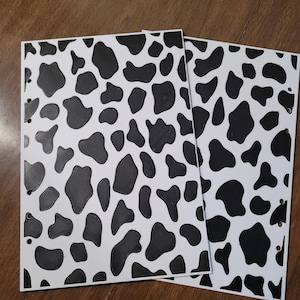 May include: Two rectangular planner inserts featuring a white background with a black cow print pattern. Each sheet has a series of circular holes punched along the left edge for binding, displaying a repeating organic spot design across the surface.