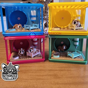 Könnte beinhalten: Vier bunte, gestapelte Hamsterkäfige. Jeder Käfig hat ein Laufrad, eine Wasserflasche und ein Miniaturbett. Die Käfige sind blau, rosa, gelb und grün. Jeder Käfig enthält eine kleine Hamsterfigur. Die Käfige stehen auf einer Holzoberfläche.