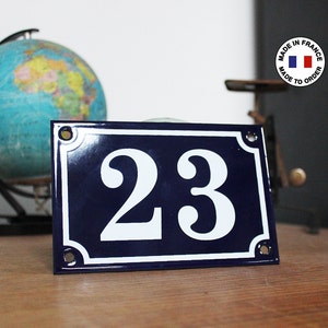 Peut inclure: Un panneau de numéro de maison en émail bleu et blanc avec le numéro 23 en blanc sur fond bleu. Le panneau a une bordure blanche et des coins arrondis.