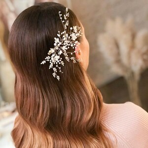 May include: A woman with long, wavy brown hair with blonde highlights. She is wearing a light pink jumper and has a delicate gold and pearl hair comb in her hair.