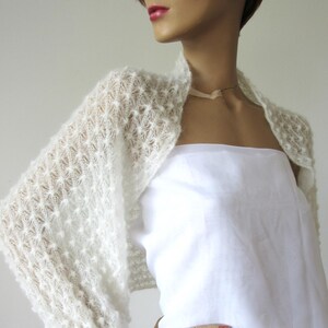 May include: A white, open-front bolero sweater with a textured, open-knit design. The sleeves are long and the sweater is worn over a white top. The bolero is displayed on a mannequin.