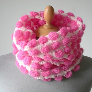 Könnte beinhalten: Ein weißer Rundschal mit einem strukturierten Muster aus erhabenen, leuchtend pinkfarbenen, kreisförmigen Noppen. Das Strickgewebe hat eine weiche, flauschige Optik und der Schal ist zu einer kreisförmigen Schlaufe gelegt, die das Punktmuster zeigt.