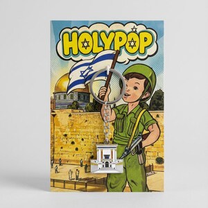 May include: A keychain featuring a detailed silver-colored building charm, attached to a metal ring. The background is a comic-style illustration of a boy in a green uniform holding an Israeli flag, with the text "HOLYPOP".