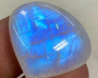 May include: A polished, cabochon-cut moonstone gemstone. The stone has a rounded shape with a smooth, translucent surface. Internal light play creates a blue and white iridescent effect. The gemstone is held by a finger.