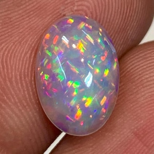 May include: A white opal gemstone with a light purple hue and iridescent flashes of red, orange, yellow, green, and blue.