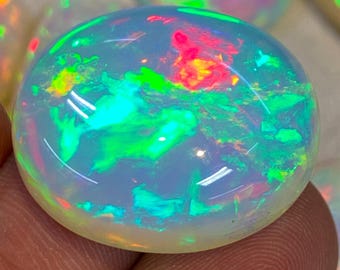 May include: A polished, oval-shaped opal gemstone. The opal displays a translucent, milky white base color with vibrant flashes of green, red, and yellow, creating a play-of-color effect. The gemstone is smooth and rounded.