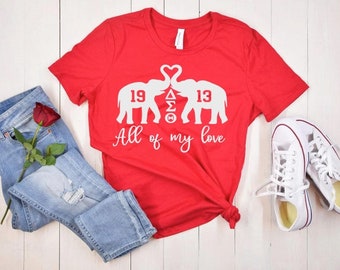 May include: Red t-shirt with white elephant silhouettes holding hearts and the text "All of my love". The elephants have the numbers "19" and "13" on them. The shirt is on a white wooden surface with a pair of white sneakers and blue jeans with a red rose.