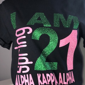 May include: Black t-shirt with the text "I AM 21 Spring Alpha Kappa Alpha" in green and pink glitter. The shirt has a crew neck and short sleeves. A pearl necklace is around the neck.