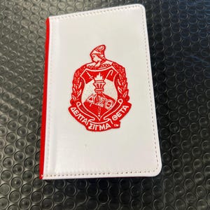 May include: White leather wallet with a red embroidered Greek fraternity crest. The crest features a torch, a shield, and the letters "DELTA SIGMA THETA".