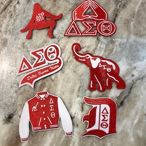 Op de afbeelding: Zeven geborduurde rode en witte patches met de Griekse letters Delta Sigma Theta. De patches tonen verschillende ontwerpen, waaronder een silhouet van een vrouw, een handgebaar, een olifant, een letter D en een varsity jack.