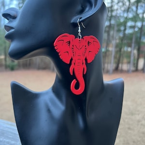 May include: Red wooden elephant earrings. The earrings are shaped like an elephant's head and have a detailed design.