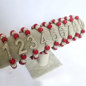 May include: Nine red and white beaded bracelets with silver number charms, numbered 1 through 9. The bracelets are displayed on a grey stand.