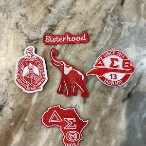 Puede incluir: Un conjunto de cinco parches bordados en rojo y blanco. El set incluye un parche con la palabra Sisterhood, un escudo de Delta Sigma Theta, una silueta de elefante, un parche circular con letras griegas y Howard 1913, y un parche con forma de África.