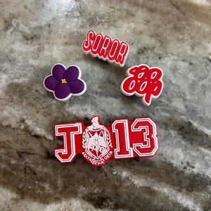 Puede incluir: Una colección de dijes para zapatos rojos y blancos. Los dijes incluyen la palabra "SOROR", una flor morada, un diseño con el número "888" y un dije con las letras "J" y "13" y letras griegas.
