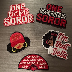 May include: Five embroidered patches with red, black, and white text and designs. The patches include phrases like "ONE DOPE SOROR" and "I'm that Delta," along with Greek letters and a fan design. The patches are on a gray surface.