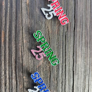 May include: Three decorative pins with the word "SPRING" and the number "25". The pins are in red, green, and blue, with the number "25" in silver. The pins are on a weathered wooden surface.