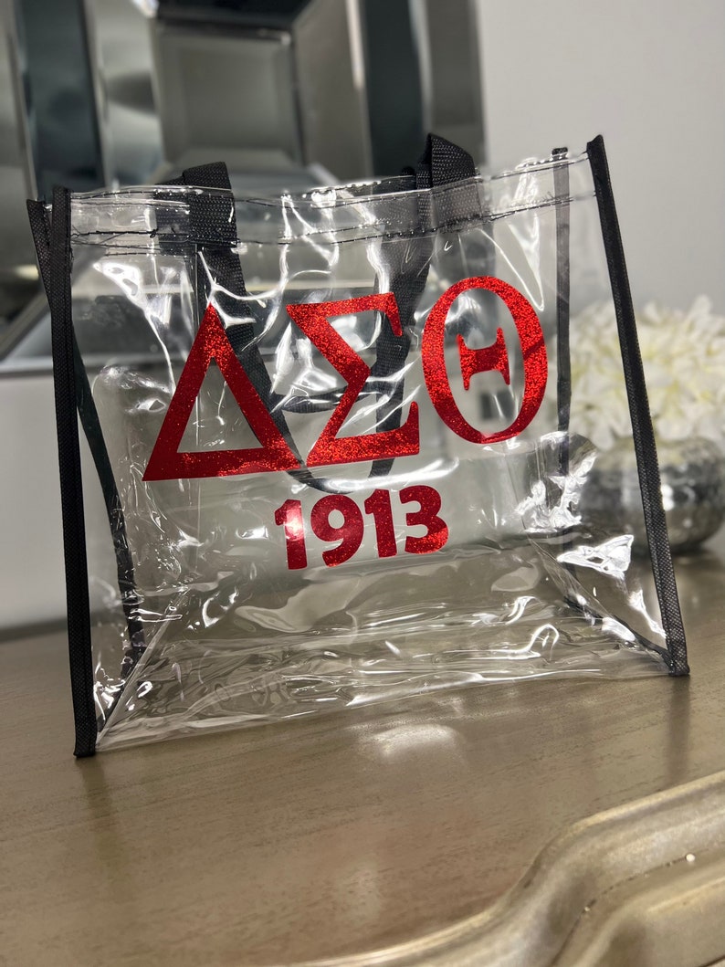 May include: Clear tote bag with black trim and handles. The bag features red glitter lettering with the Greek letters Delta, Sigma, and Theta, along with the number 1913. The bag is made of a transparent material.