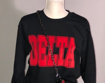 May include: Black sweatshirt with a red "DELTA" graphic on the front. A black chain strap purse is worn over the sweatshirt.