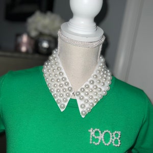 May include: A green short-sleeved shirt with a white collar adorned with faux pearls. The collar is attached to a mannequin head. The number "1908" is spelled out in pearls on the shirt.